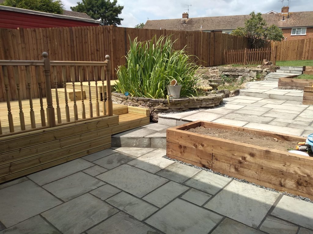 A wide view of a paved garden with multiple features, planters, decking and plants etc