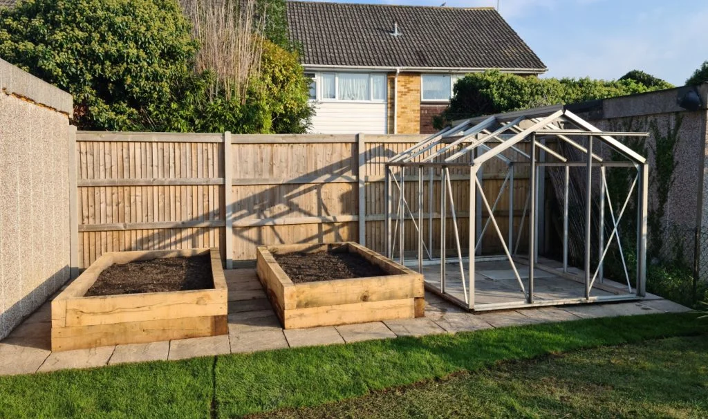 Artificial grass installation in a garden with paving, planters and greenhouse frame
